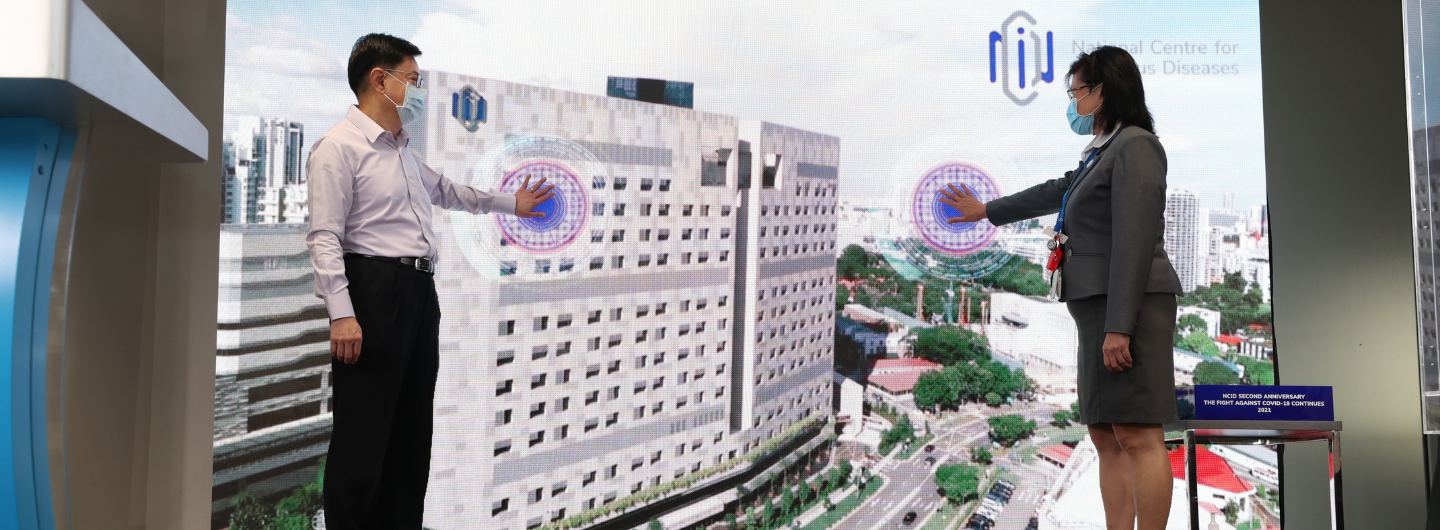 Two masked people point at a large screen showing NCID building and logo.
